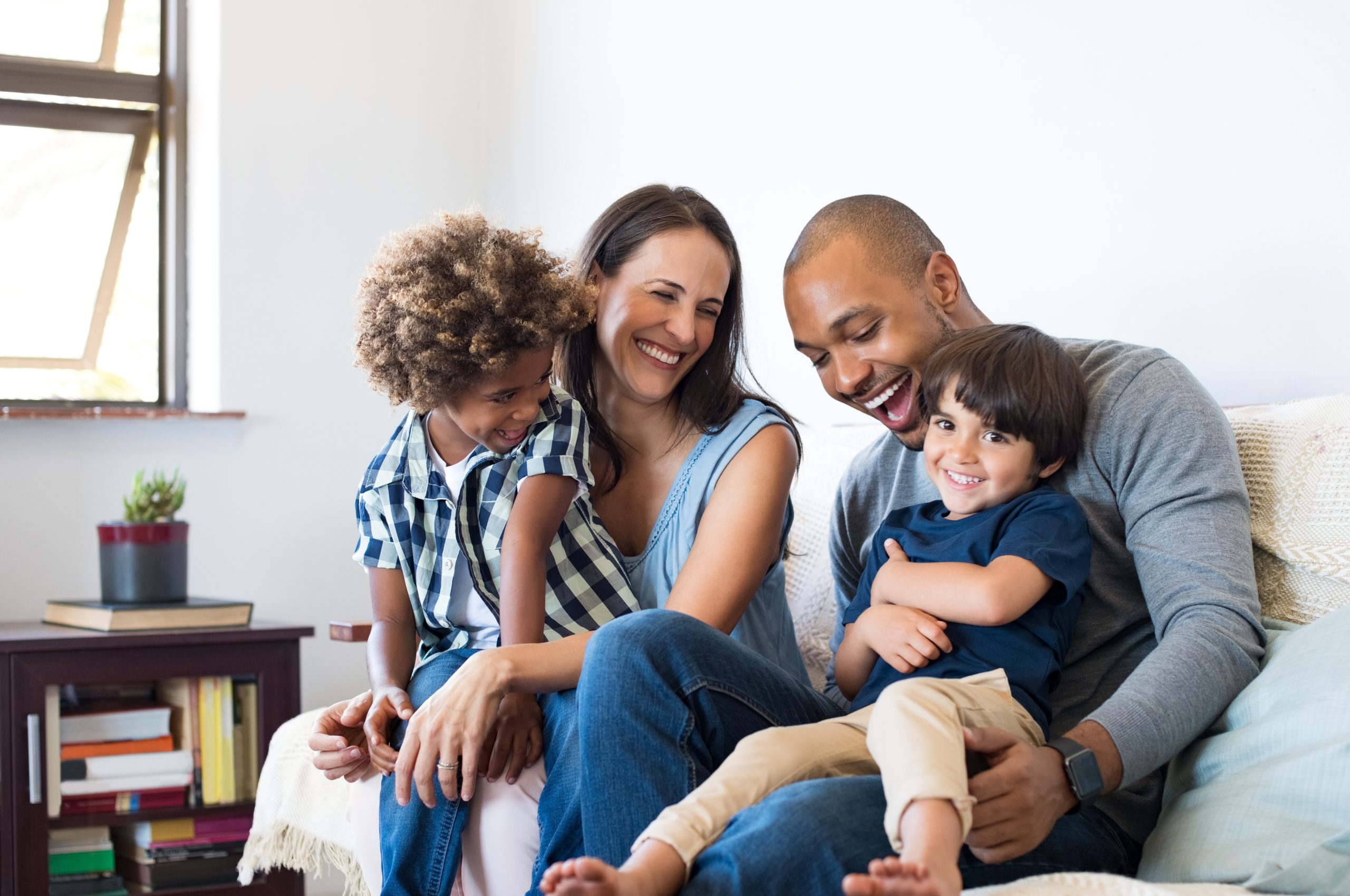 A happy family of four sits together on a couch, smiling and laughing. The parents are in the middle, with their two young children on either side, all enjoying time together in a cozy, well-lit room.