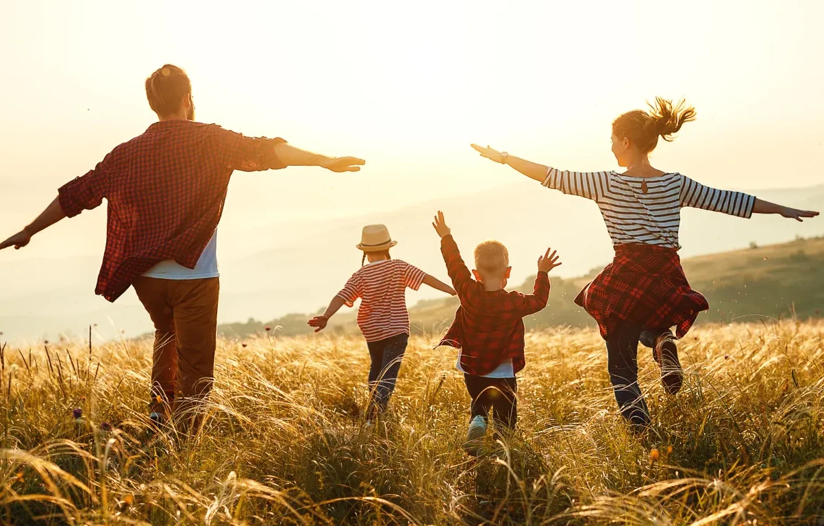 A family of four, two adults and two children, run joyfully through a sunlit field with arms outstretched, wearing casual clothes and surrounded by tall grass at sunset.