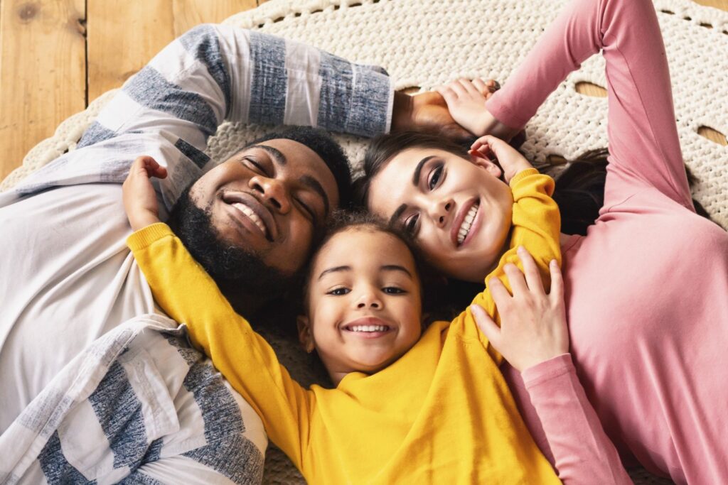 A happy family of three lies on a rug, smiling up at the camera. The child, in a yellow shirt, is in the middle, hugging both adults, who are lying on either side, creating a warm and joyful scene.