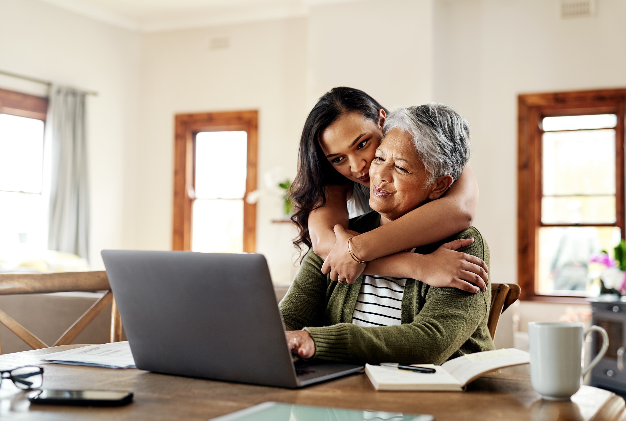 A young woman hugs an older woman from behind as the older woman uses a laptop at a table. Both are smiling, and the scene is set in a bright, cozy living room with books and papers on the table.