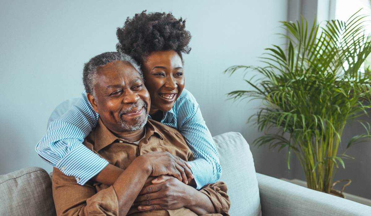 A smiling young woman hugs an older man from behind as they sit on a couch in a bright living room, with a large green plant in the background.