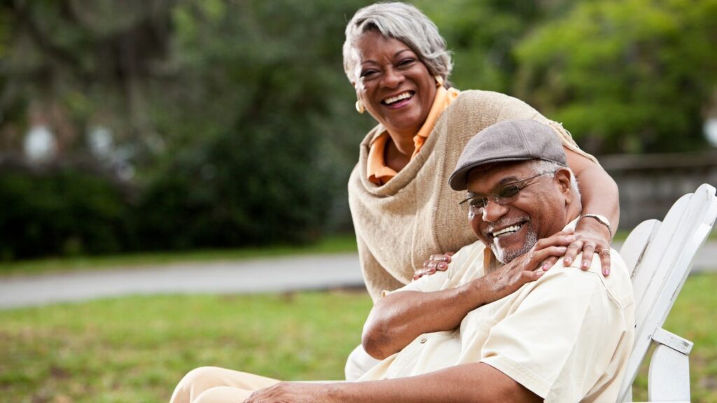 An older couple sits together outdoors on a white bench, smiling and laughing. The woman stands behind the man with her hands on his shoulders; both look happy and relaxed in a green park setting.