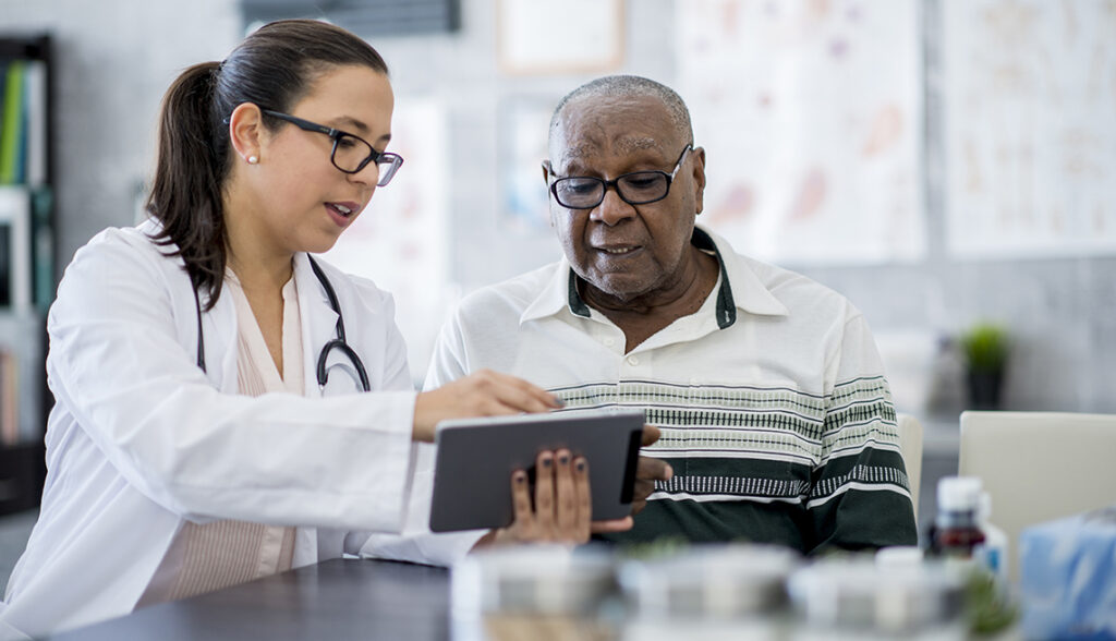 A senior man of African-American descent is indoors in a hospital room. He is watching his female doctor using a tablet computer.