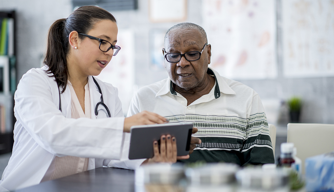 A senior man of African-American descent is indoors in a hospital room. He is watching his female doctor using a tablet computer.