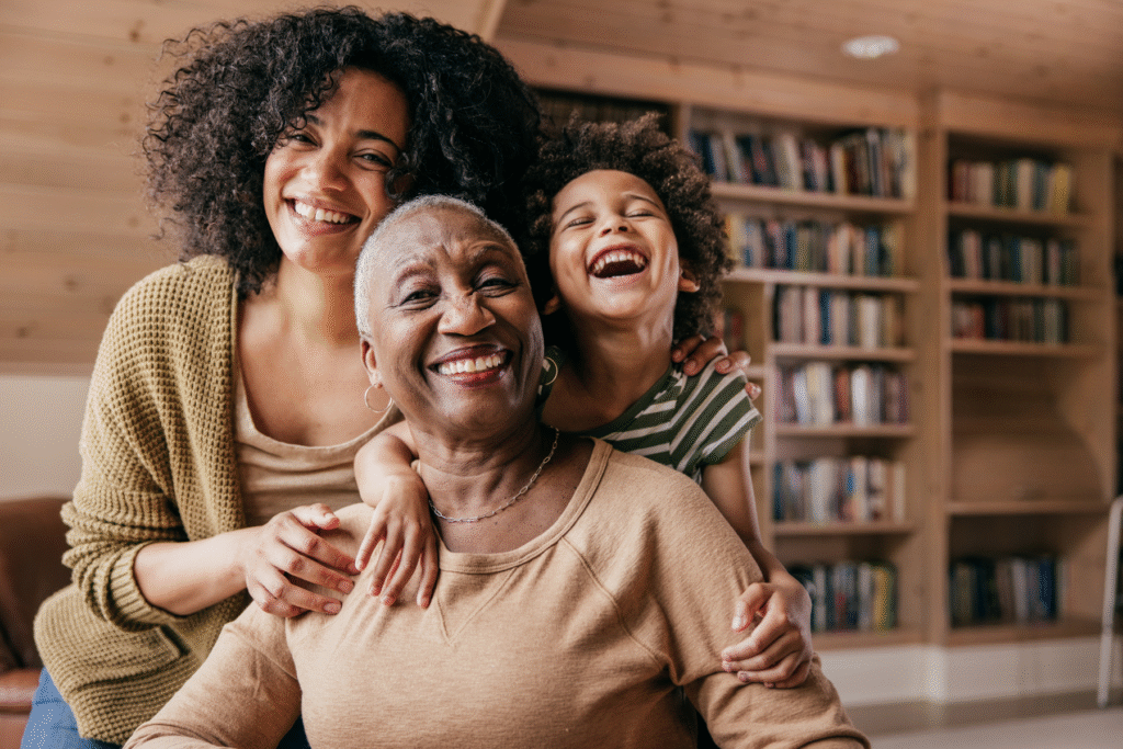 Three generations—an older woman, an adult woman, and a young child—smiling and hugging each other in a cozy living room with bookshelves in the background.