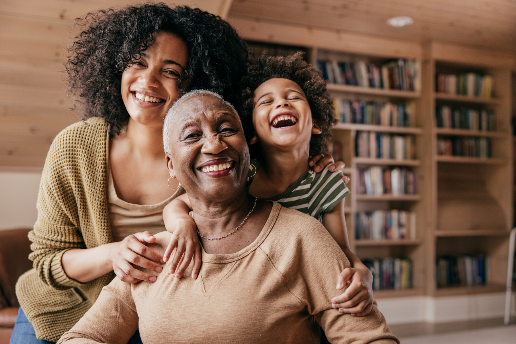 Three generations—an older woman, an adult woman, and a young child—smiling and hugging each other in a cozy living room with bookshelves in the background.