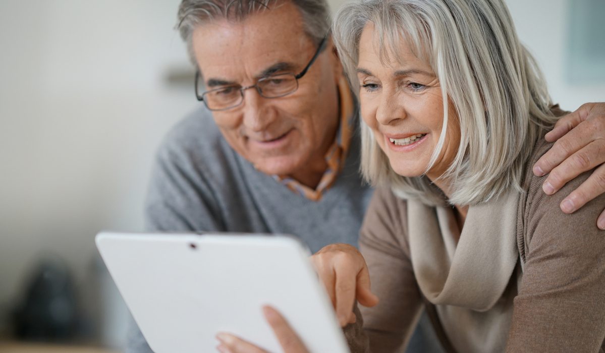 An older couple sits closely together, smiling and looking at a tablet screen. The man wears glasses and a gray sweater, while the woman has gray hair and wears a beige scarf and top. They appear happy and engaged with the device.