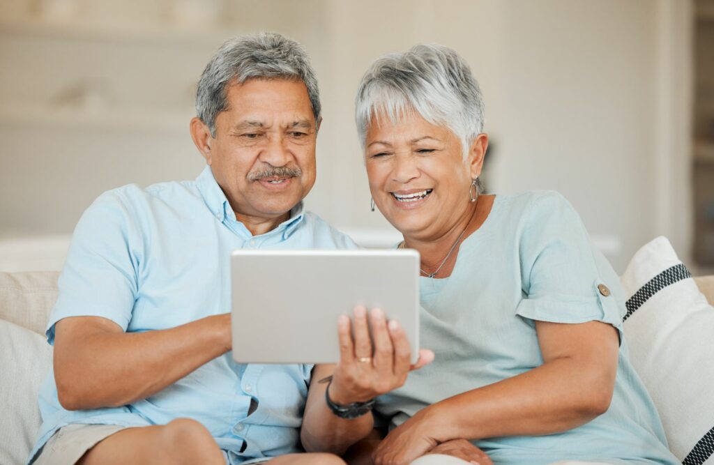 An older couple sits closely together on a couch, smiling and looking at a tablet device they are holding in their hands. The background is bright and softly lit, creating a warm, comfortable atmosphere.