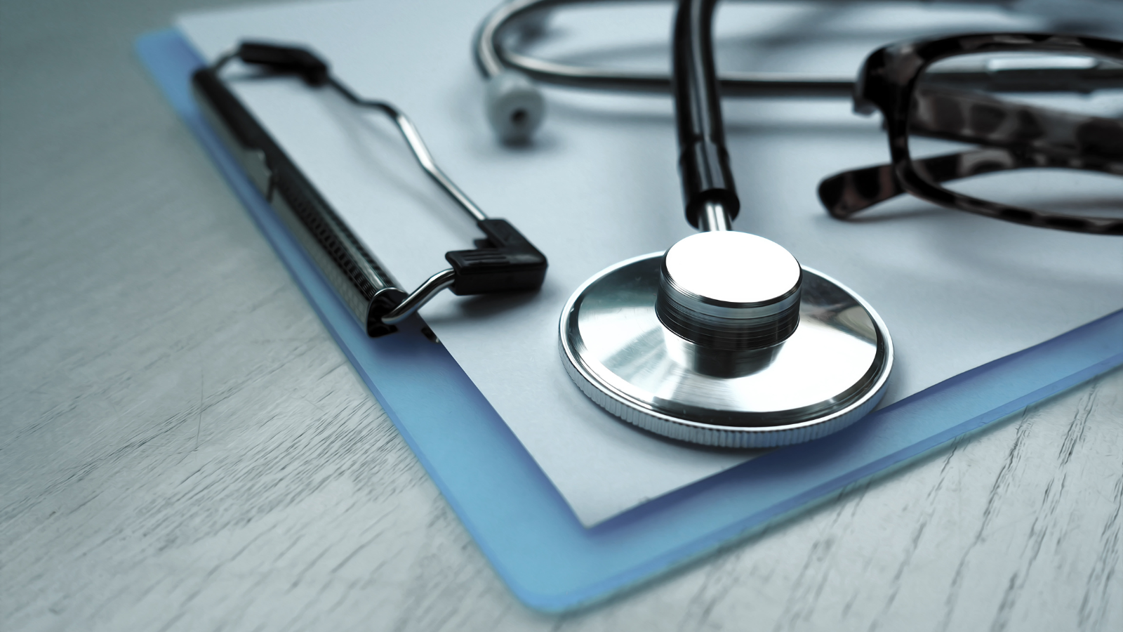 Close-up of a stethoscope, clipboard, and eyeglasses resting on a stack of papers with a blue folder, placed on a light wooden surface.