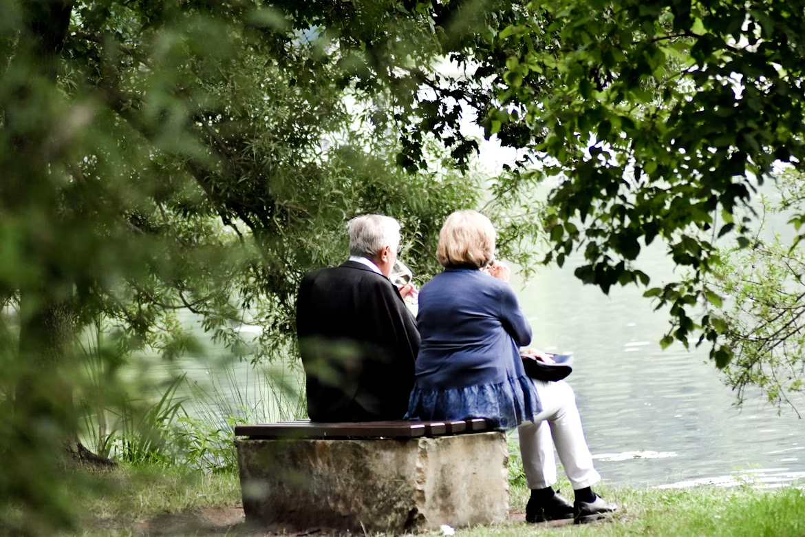 Two older adults sit together on a bench by a lake, surrounded by green trees and foliage, peacefully enjoying the tranquil outdoor setting.
