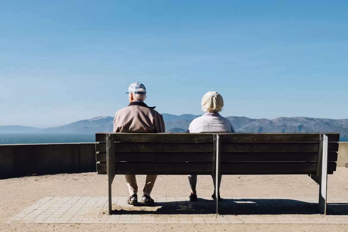 Two older adults sit side by side on a bench, facing the ocean and distant mountains under a clear blue sky.