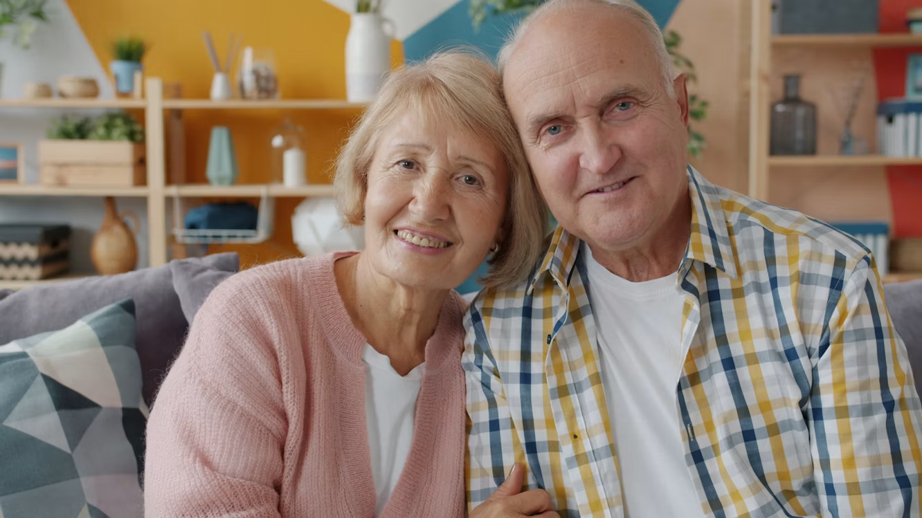 An elderly couple sits close together on a couch, smiling warmly at the camera in a cozy, colorful living room.