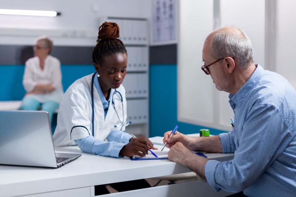 A doctor in a white coat and an older man with glasses writing on paperwork together at a clinic desk
