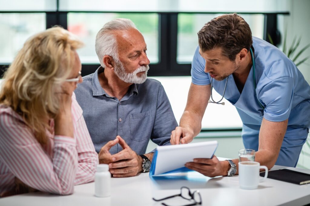 A doctor in blue scrubs pointing to a folder of documents while an older man and woman look on at a table