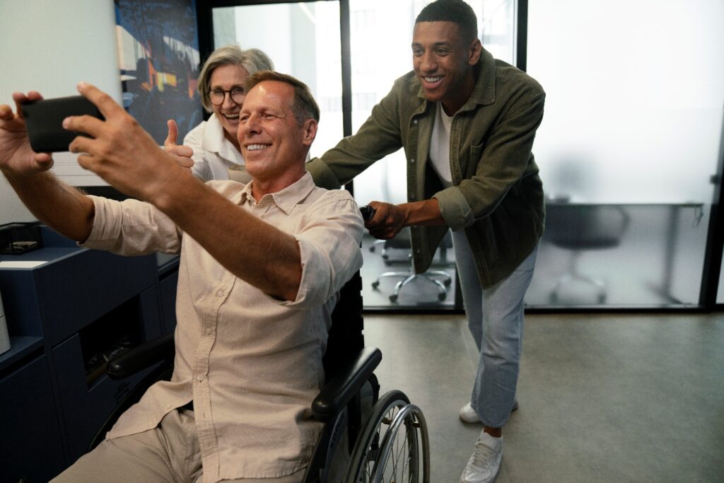 A man in a wheelchair takes a selfie in an office while a smiling older woman gives a thumbs up and a younger man stands behind him