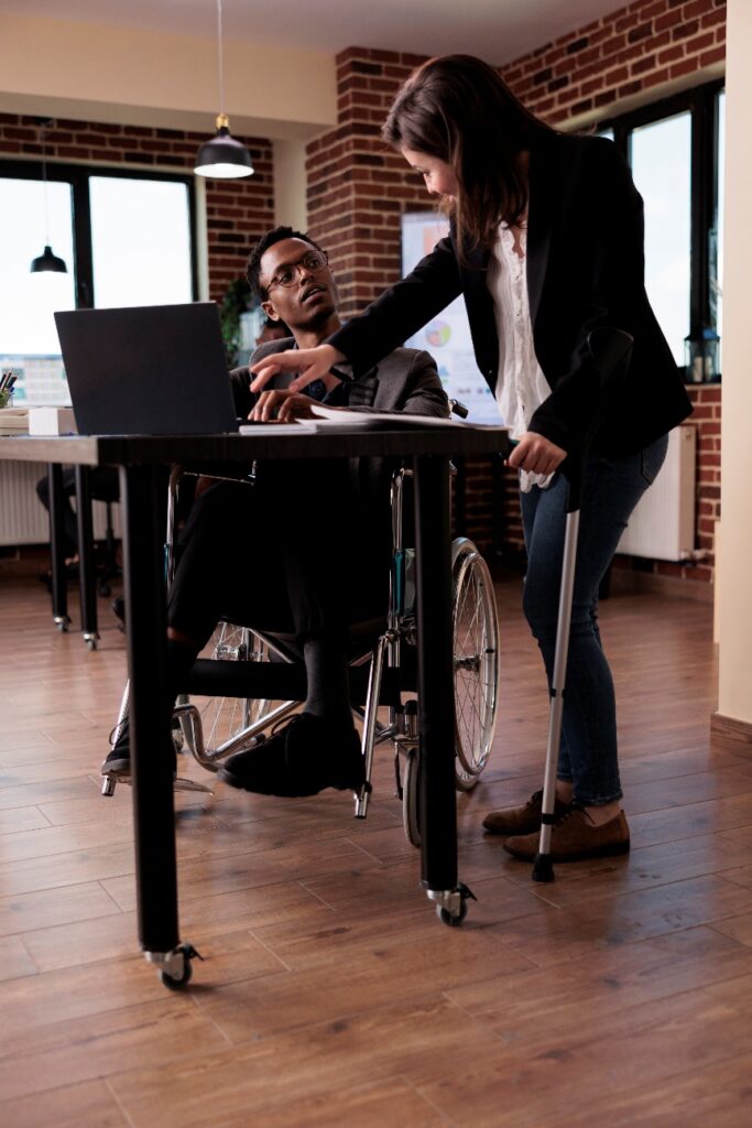 A man in a wheelchair at a desk looking up at a woman using a crutch who points at his laptop screen in a brick-walled office