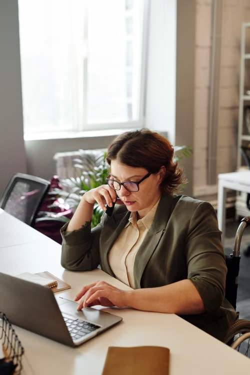 A woman in a wheelchair working on a laptop while talking on a phone at a home office